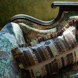 A decorative throw pillow with a book spine pattern and ruffled trim, resting on an ornate vintage-style upholstered armchair, with fabrics and wallpaper from the Disney Belle Collection.
