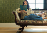 A woman reclining on an ornate Disney's Belle floral chaise lounge and reading a teal book in a room with green Belle damask wallpaper and a window.