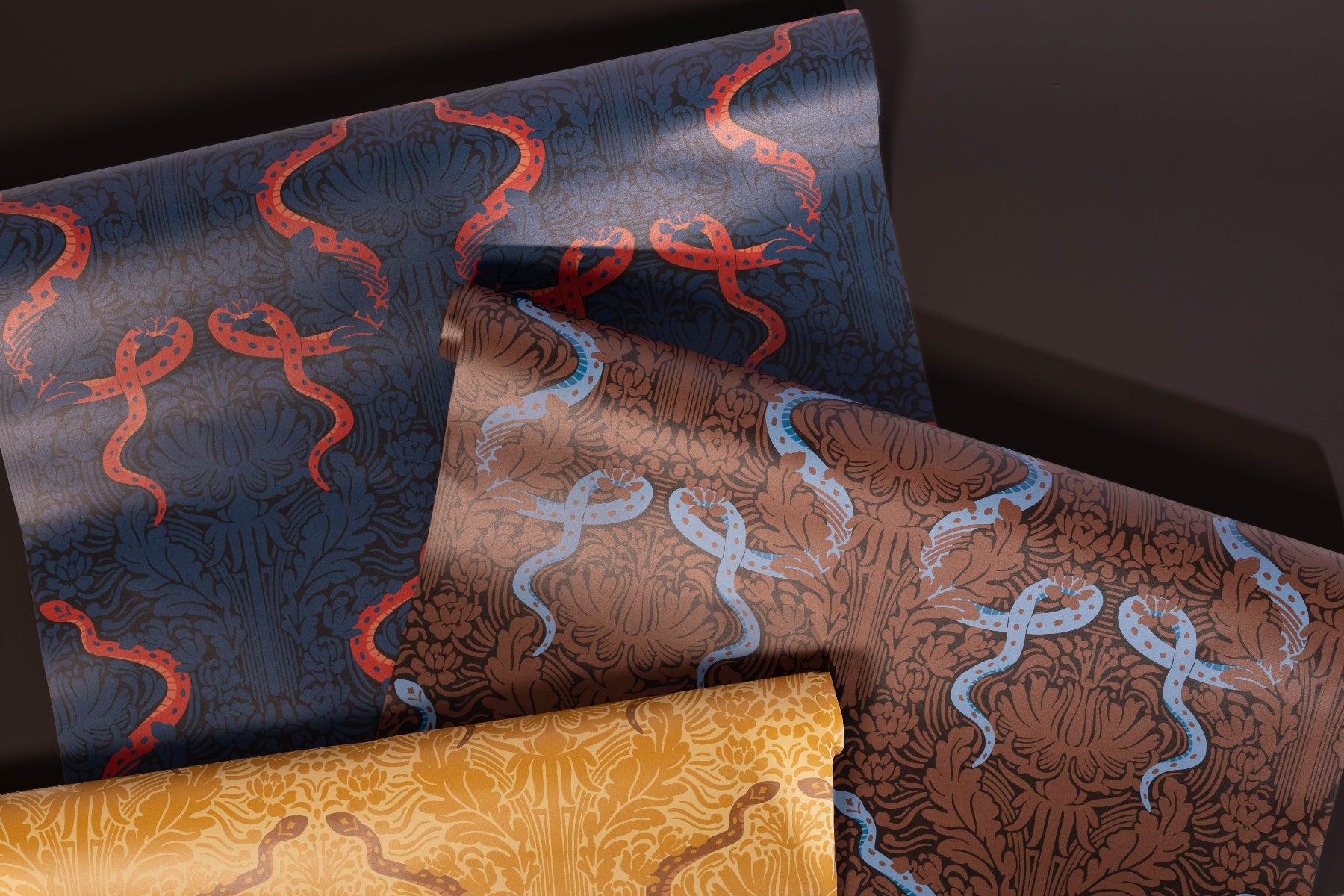 Three rolls of patterned wallpaper displayed diagonally, featuring a dark blue roll with red snakes, a brown roll with light blue snakes, and a gold roll with dark brown snakes.