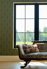 A large black-framed multi-pane window set in a room with green damask Disney's Belle-inspired wallpaper, light wood flooring, and a patterned upholstered chaise lounge.