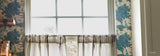 A set of sheer white tiered curtain panels with gray and white striped valances hanging in front of a bright, overexposed window. The room features blue and green Disney Tiana toile wallpaper on the sides.