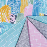 A close-up of folded Spider-Man fabric showing multiple printed patterns and colors: blue cityscape buildings, pink and yellow buildings, white brick lines, gray spiderweb lines, and a pink patterned section.