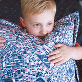 A young boy with is lying down and holding a large decorative pillow against his face. The pillow features a geometric Spider-Man pattern of navy blue, white, and coral red lines.