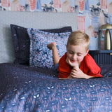 A young boy with lies on his stomach on a bed. The bed is made with navy blue Spider-Man bedding featuring a white and red abstract pattern, with matching patterned pillows and a gray headboard.
