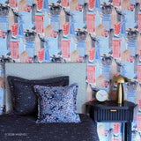 A styled bedroom scene featuring a bold cityscape patterned Spider-Man wallpaper in blue, red, and gray. In the foreground, a bed with dark patterned Spider-Man bedding, a gray upholstered headboard, a black nightstand with a gold lamp, and decorative pillows are arranged.