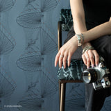 A person seated on a dark wooden chair against a Spider-Man blue-gray wallpaper with a repeating pattern. The person is wearing a black top and dark pants, holding a vintage silver camera, with bracelets and rings visible on the hands.