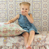 A young girl with blonde pigtails sits on a Disney's Rapunzel-inspired floral-patterned sofa, wearing a denim blue romper dress. She is resting her hand on a matching floral pillow in front of a patterned wallpaper background.