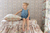A young girl with blonde hair in a ponytail sits on a Disney Rapunzel-inspired floral-patterned bed, wearing a blue denim romper. She is smiling and looking to the side.