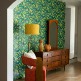 A styled interior room featuring a bold botanical Spider-Man wallpaper in teal, green, and yellow on an arched wall. In front of the wall is a mid-century modern wooden dresser with a matching arched mirror, a gold floor lamp, and two decorative vases.