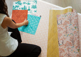 A person kneels on a light pink Spider-Man wallpaper, handling several rolled and flat sheets of other Spider-Man wallpaper. Visible designs include teal with white toile-style motifs, rust orange with repeating scallop shapes, a yellow-gold scallop pattern, and a blush pink botanical print with blue accents.